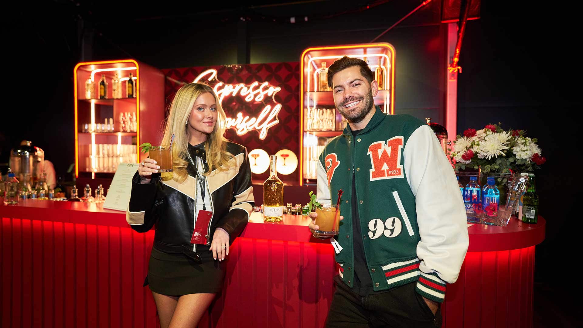 A woman and man in leaning against a red bartop, whilst holding drinks in their hands posing for a photo