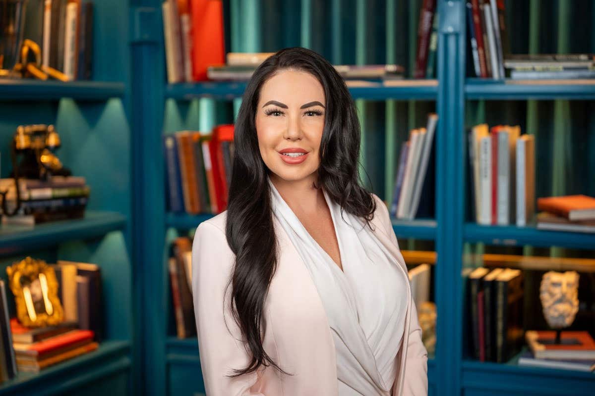Woman with long dark hair wearing a light pink blazer and white blouse, smiling in front of blue bookshelves filled with books and decor.