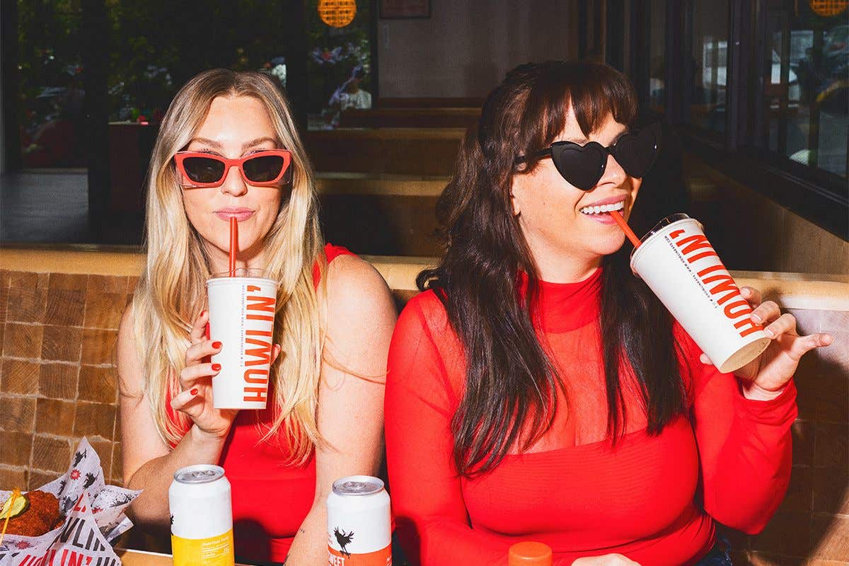 Two women sitting in a booth with red shirts and sunglasses on sipping on straws from their to-go cups. 