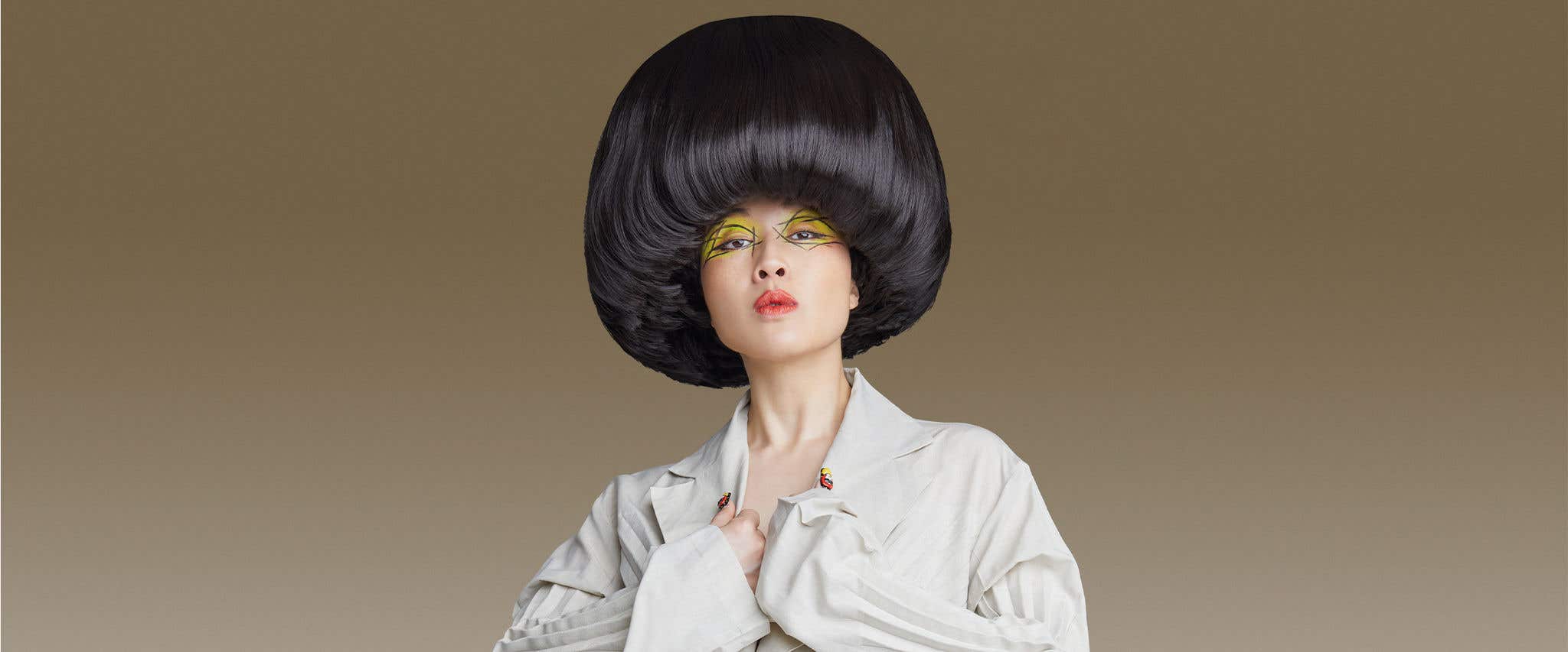 Atsuko Okatsuka in white shirt with big bowl cut hair standing in front of beige background looking at the camera.