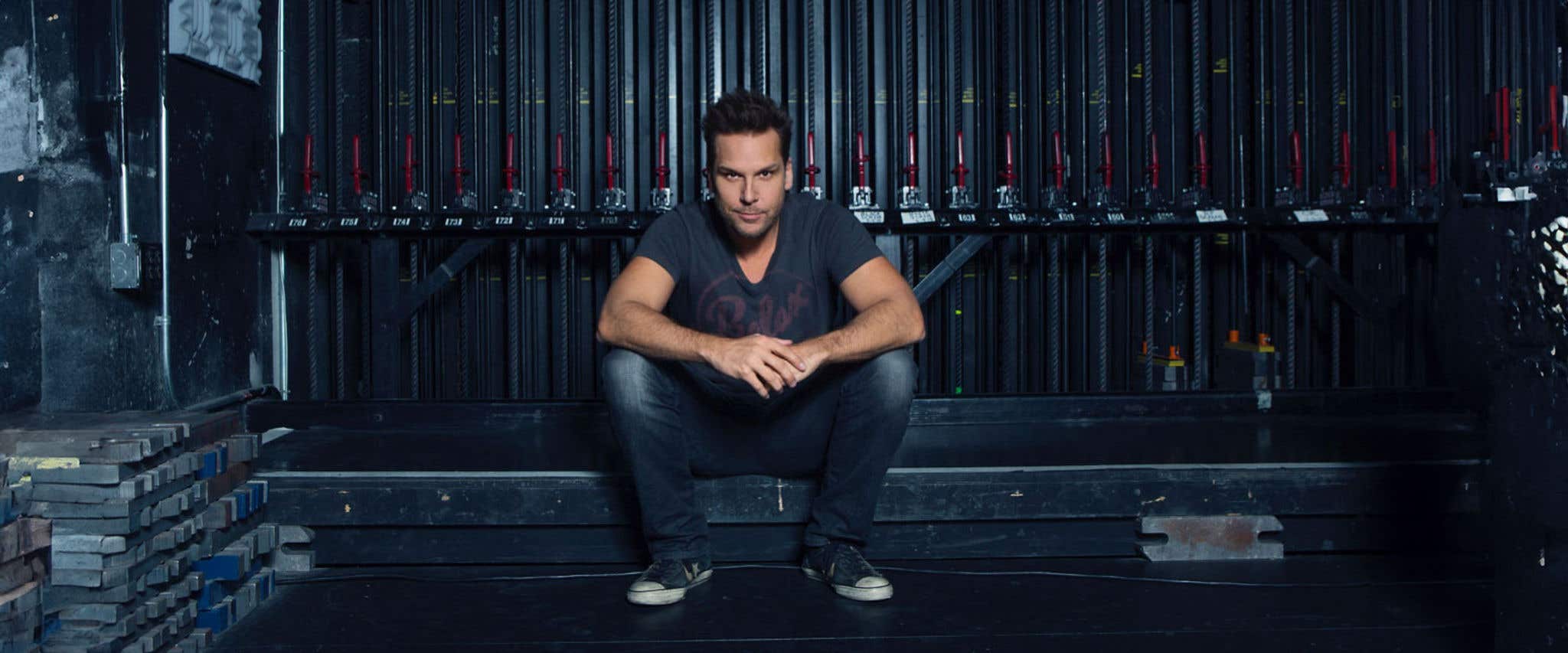 Dane Cook sitting in front of ropes and levers in jeans and a t-shirt staring directly at the camera.