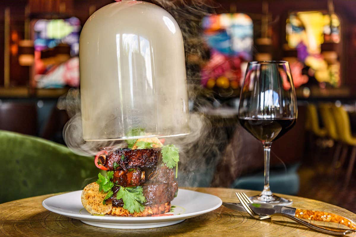 A steak dish with smoke under a glass dome beside a glass of red wine on a wooden table in a colorful restaurant.