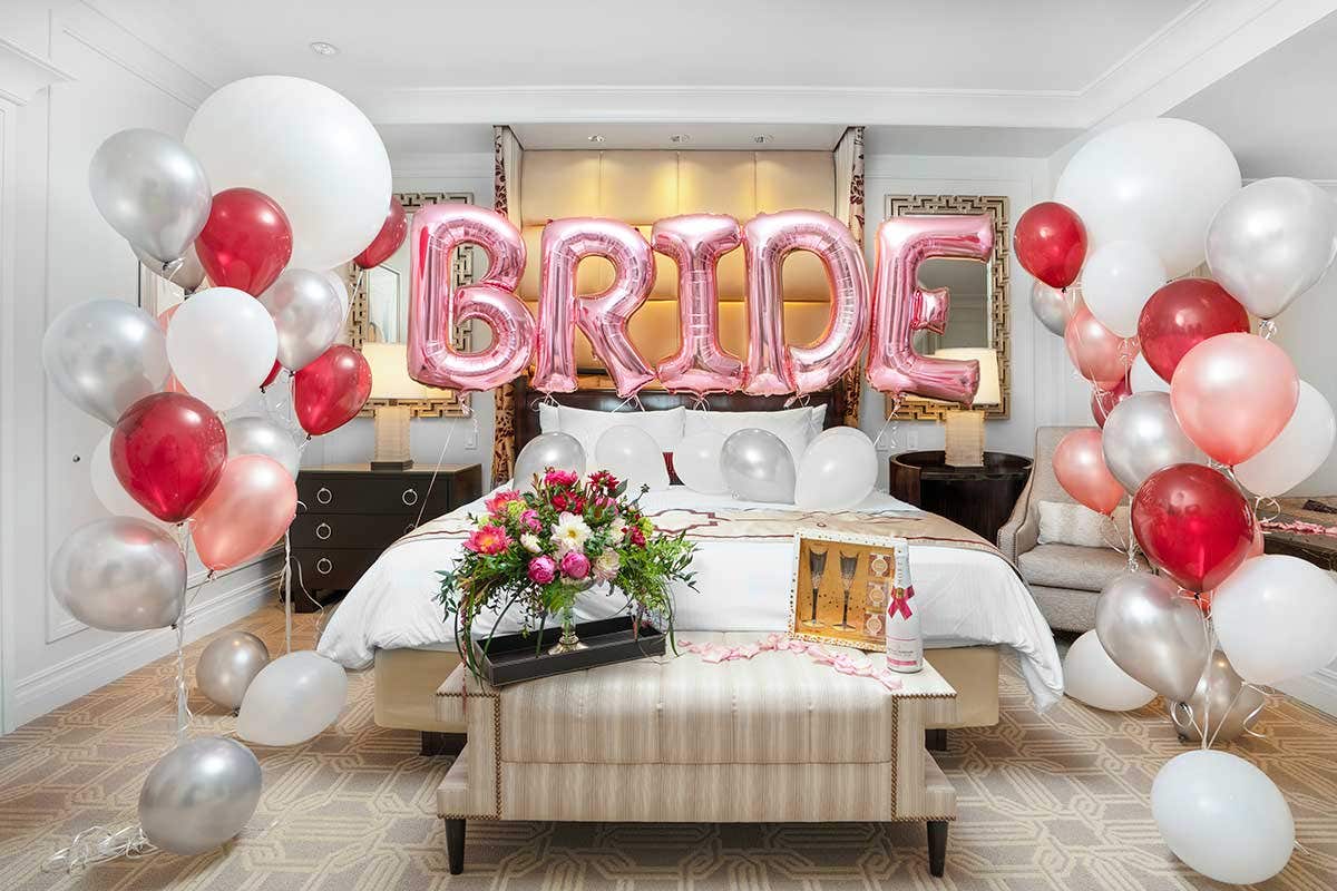 Hotel room decorated with balloons, flowers, and large pink "BRIDE" balloons above the bed.