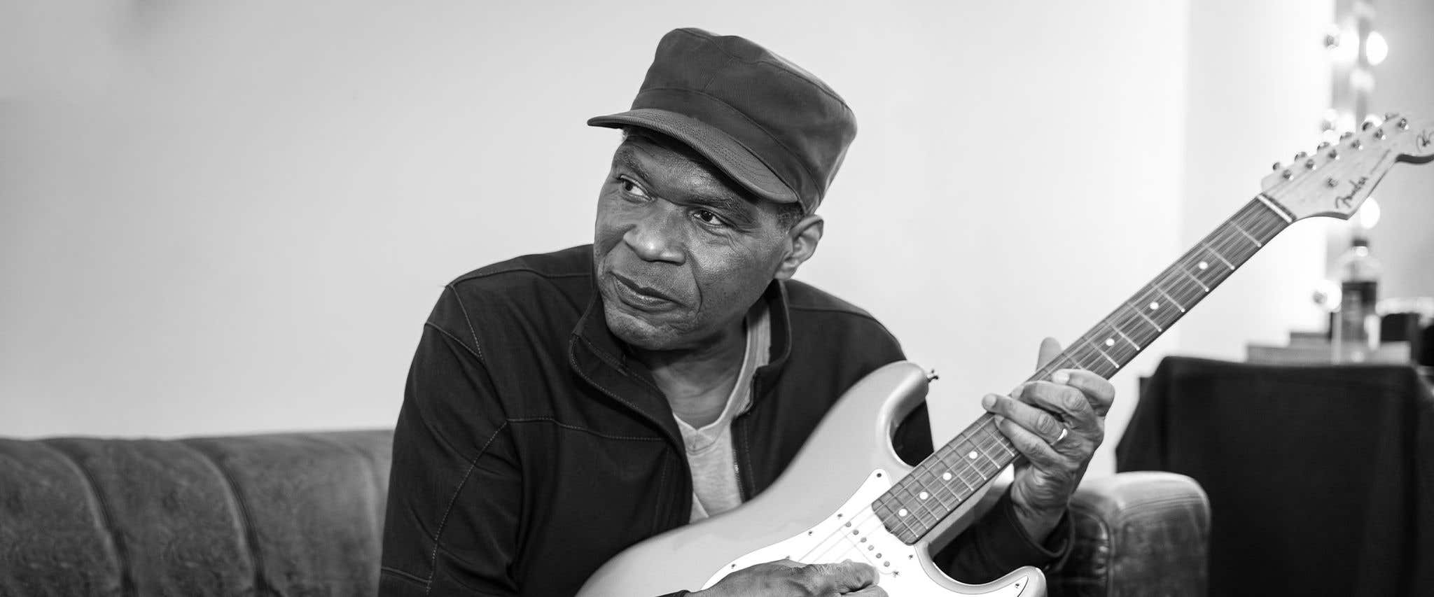 A black and white photo of Robert Cray holding a guitar while sitting on a couch