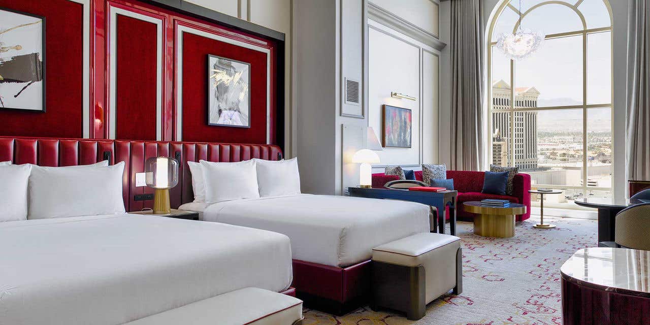A hotel room with two queen beds, an armchair, a red curved couch, a gold coffee table, and a large arched window.
