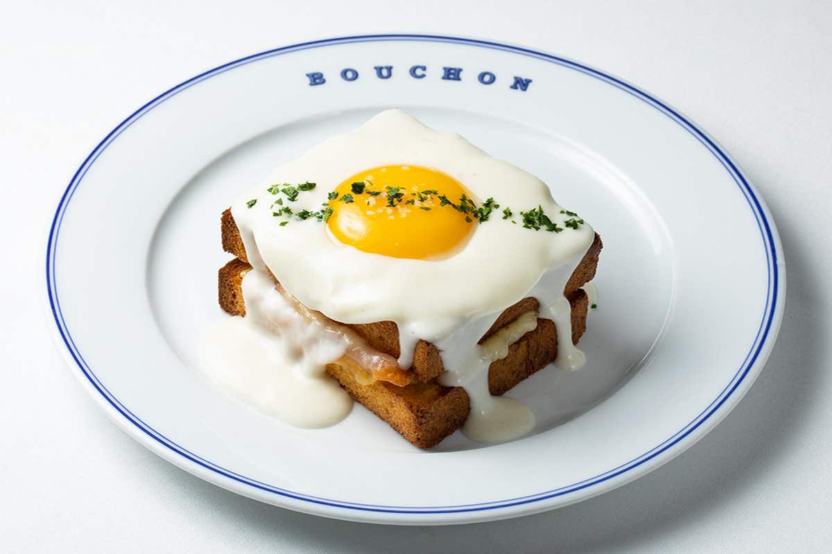 A sunny-side-up egg on a stack of cheesy, creamy toast with greens, served on a plate labeled "Bouchon".