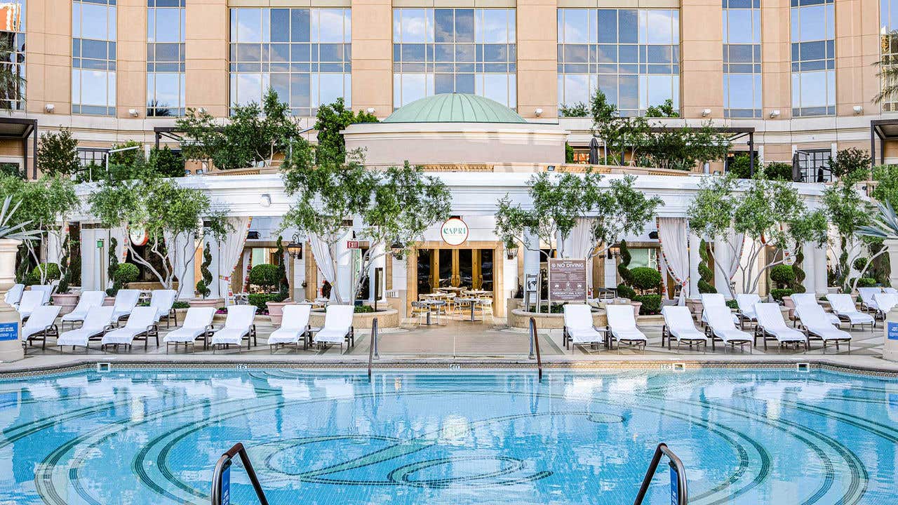A pool with lounge chairs on the edge with an open patio restaurant behind the pool.