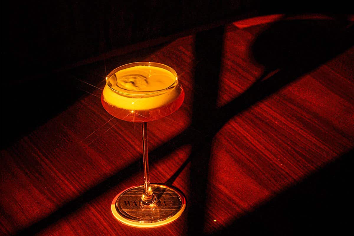 A cocktail glass with a frothy yellow liquid on a red-wooden surface, highlighted by dramatic sunlight.