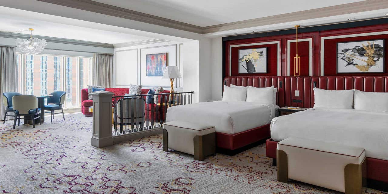 A hotel room with two queen beds, a sitting area, and large windows.