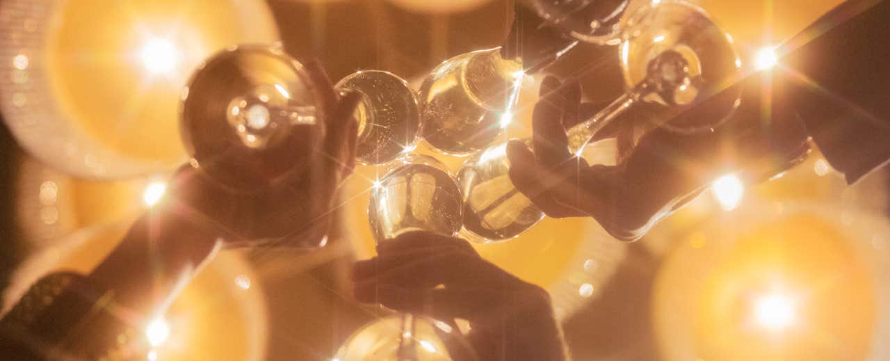 Hands holding champagne flutes in a toast, with glowing lights in the background creating a festive atmosphere.