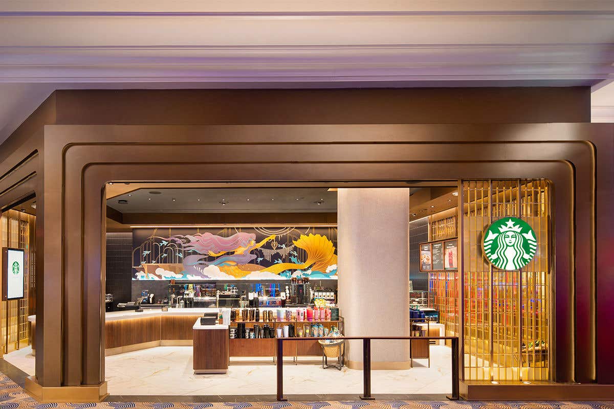 Coffee Shop with a colorful image of a mermaid on the back wall above a coffee station including, spouts, and teas. Coffee cups lined up on an island in the middle of the shop with a green Starbucks circle logo which has a green background with a white mermaid with a crown. 