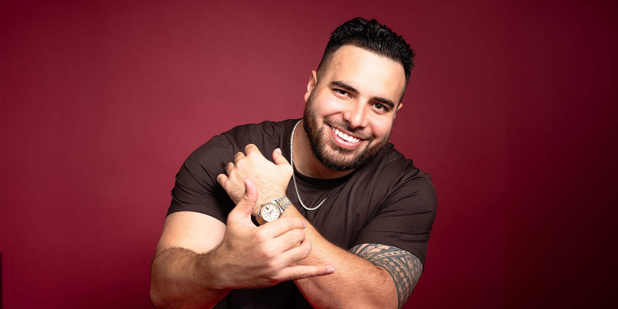Comedian Tumua smiling, resting on one arm, set against a red background.