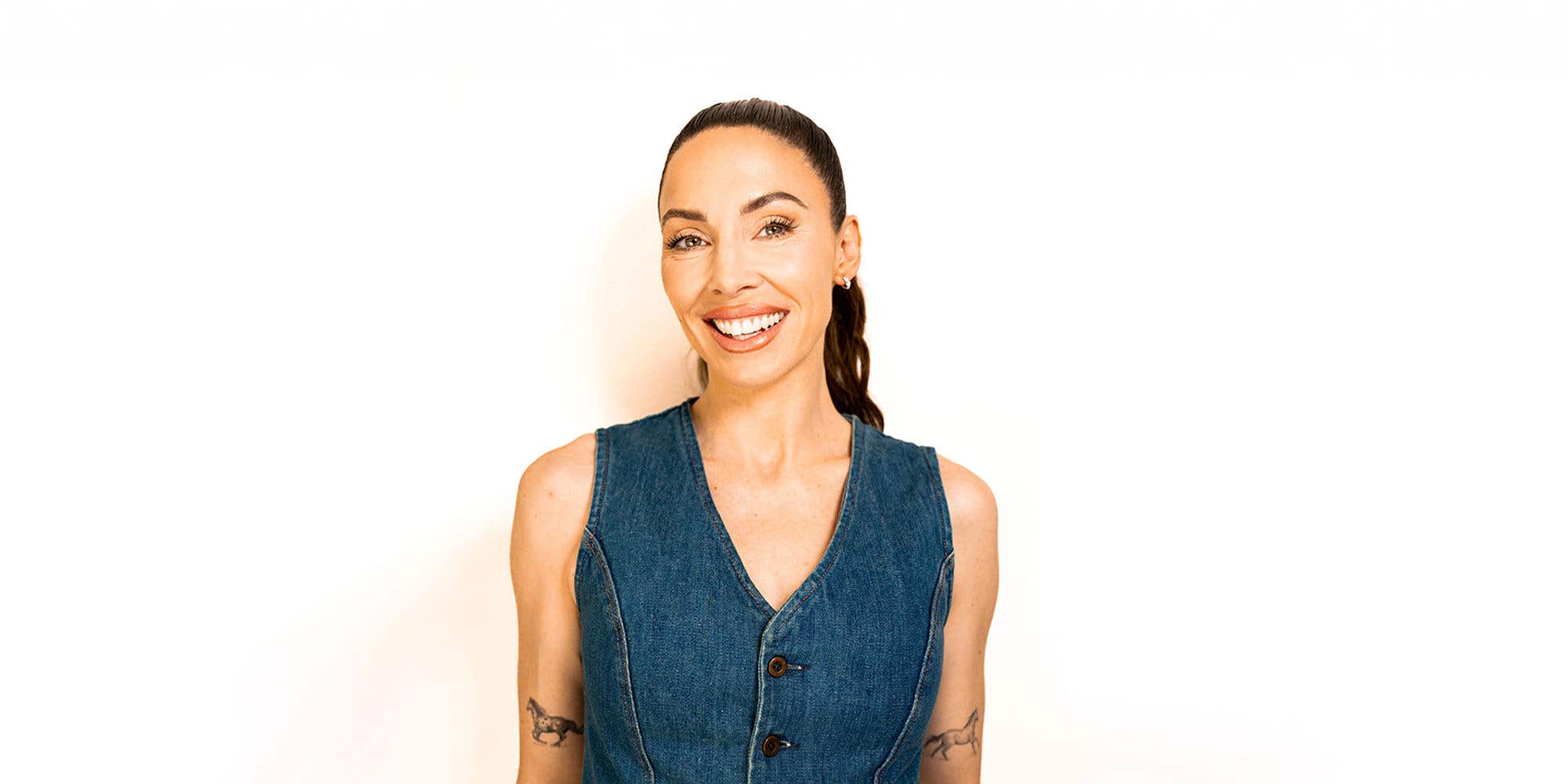 Comedian Whitney Cummings standing against a white background, wearing a sleeveless denim top.