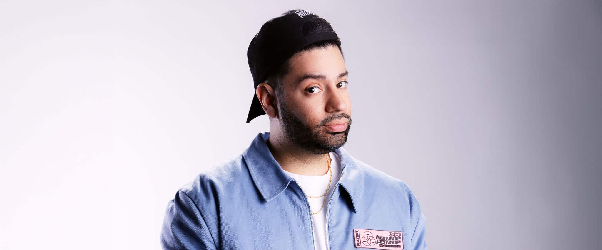 Martin Amini in black backward facing hat, white shirt and blue jacket standing in front of neutral gray background looking directly at camera.  