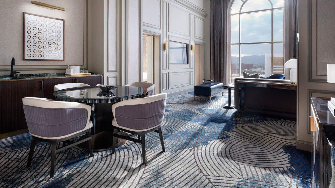 A hotel living room with a dining table with four chairs, a wet bar, TV, bench, couch, and tall arched window with a view of the Las Vegas Strip.