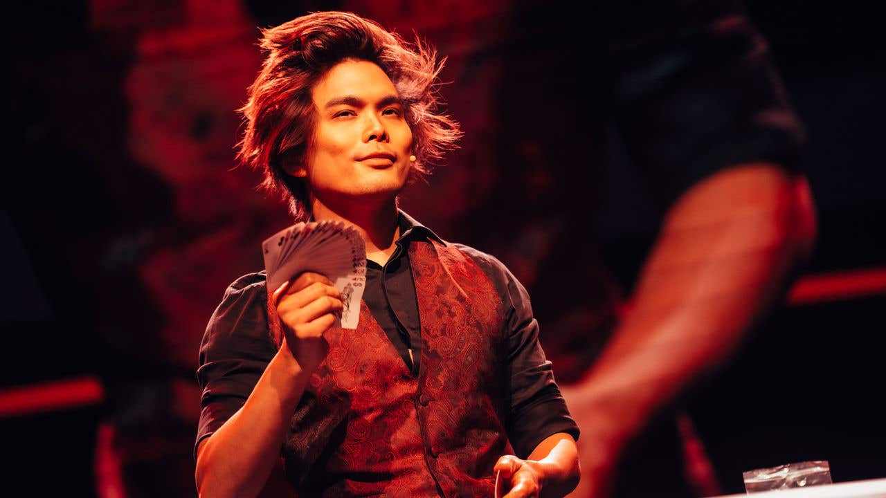 Shin Lim in a burgundy vest and black shirt smiling while holding a fanned out deck of cards. 