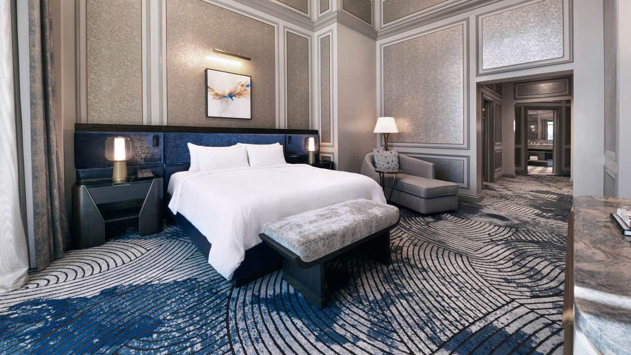 A hotel bedroom with a king bed, bench, chaise lounge, high ceilings, and a connected bathroom.