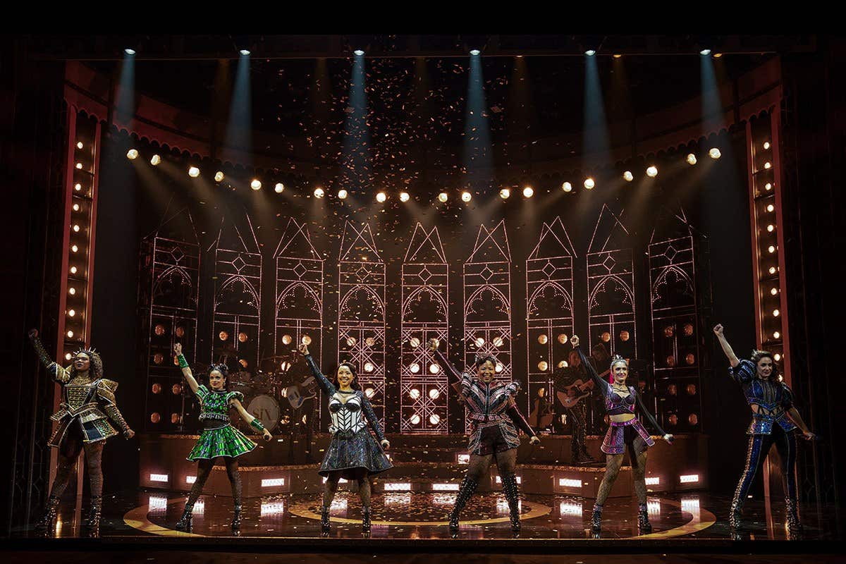 Six performers in colorful, ornate costumes on a stage with a flashy, medieval backdrop, raising their arms triumphantly.