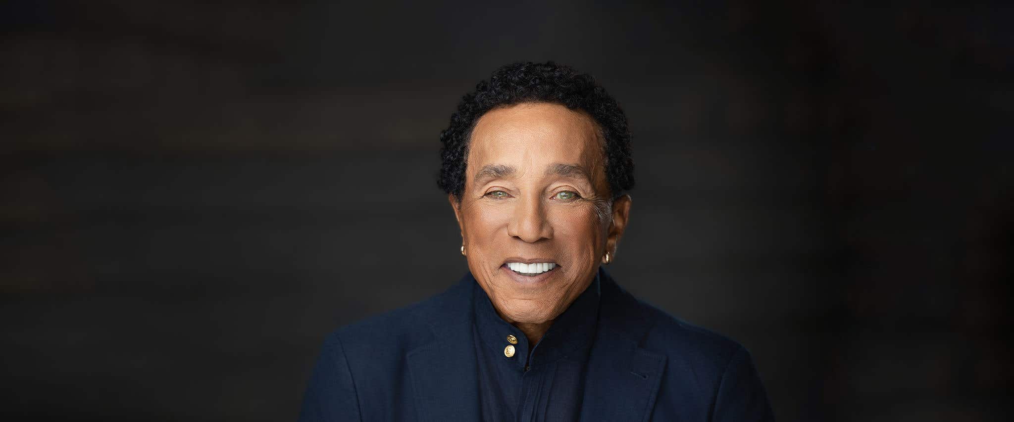 Smiling person with short curly hair, wearing a dark blue suit against a dark gray background.