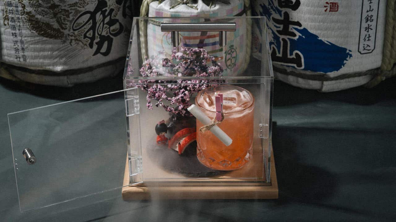 A cocktail displayed inside a clear case with decorative cherry blossoms and a miniature bridge.