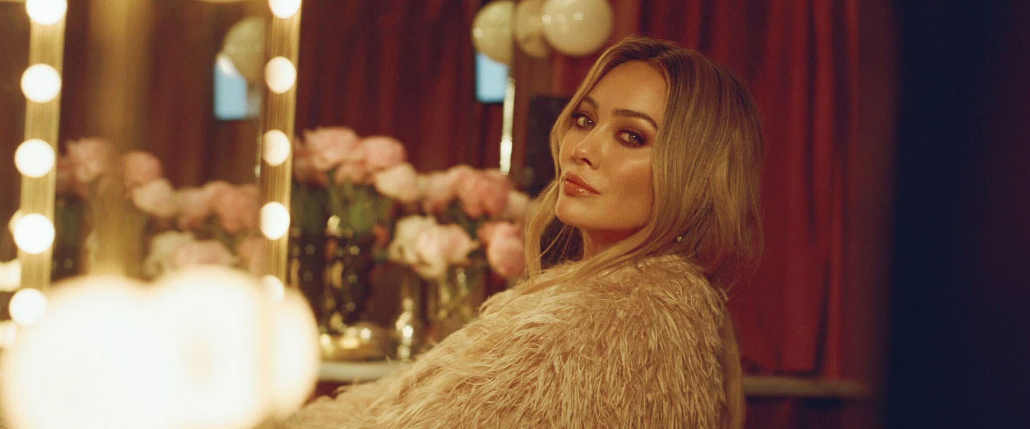 Hilary Duff sitting on clear seat in a fur coat in front of a vanity table looking towards the camera. 