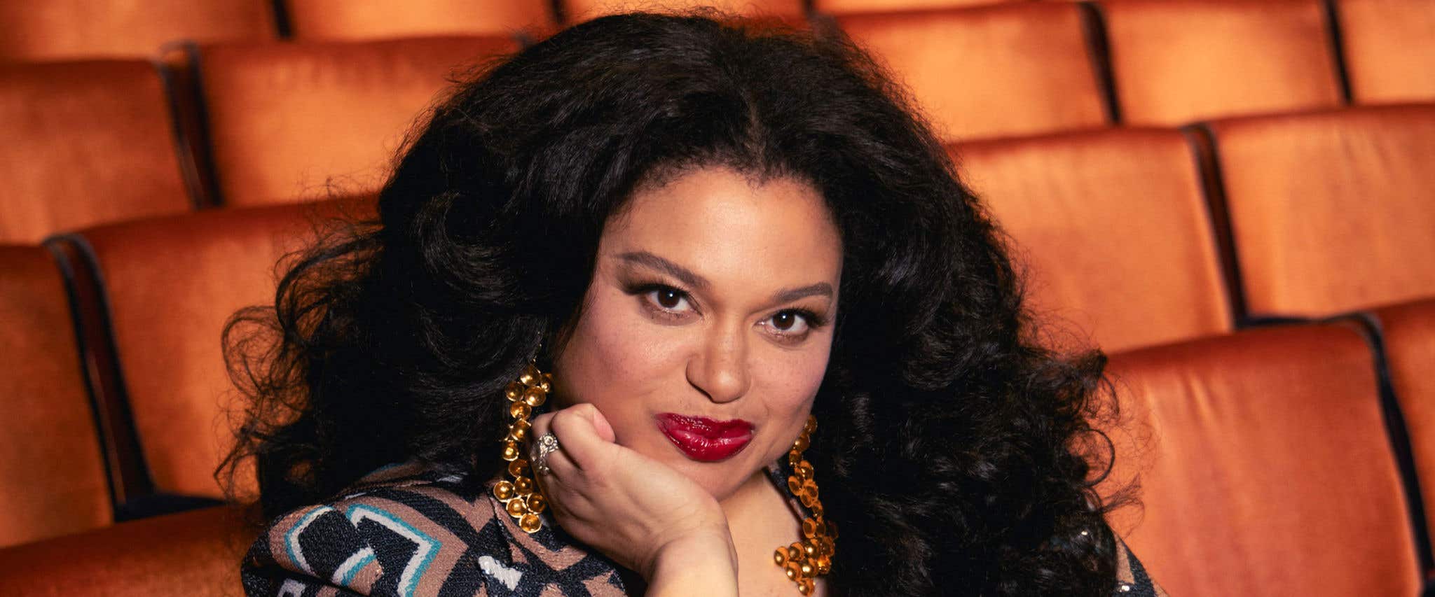 Michelle Buteau wearing a long-sleeved top, sitting in a theatre seat resting her arm on the seat in front of her and surrounded by empty theatre seats.