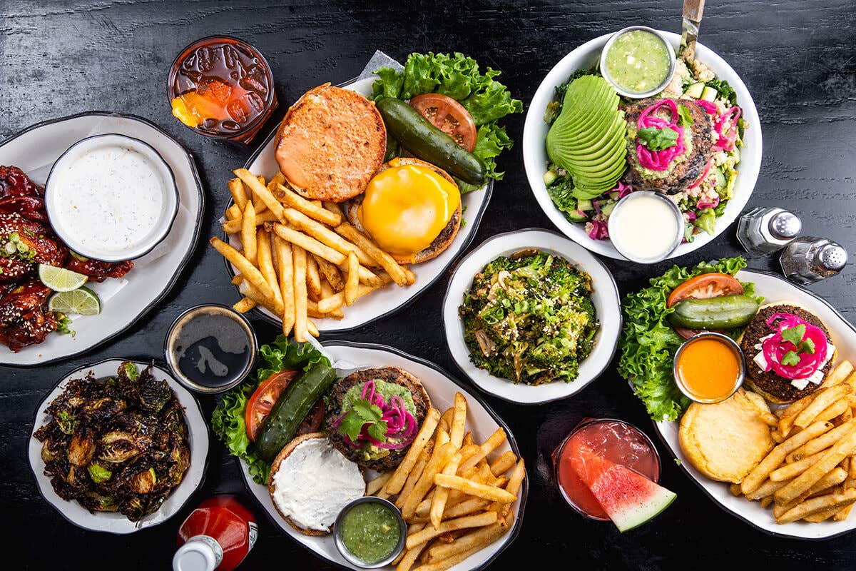 A variety of dishes including burgers with fries, salads, roasted veggies, sauces, and drinks on a dark table.