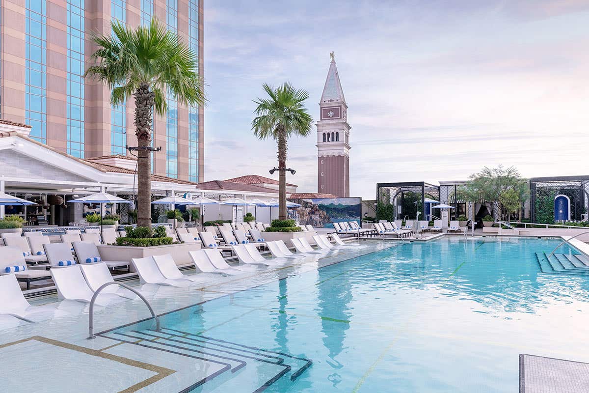 Outdoor pool area with lounge chairs, palm trees, and a tall clock tower in the background, under a clear sky.