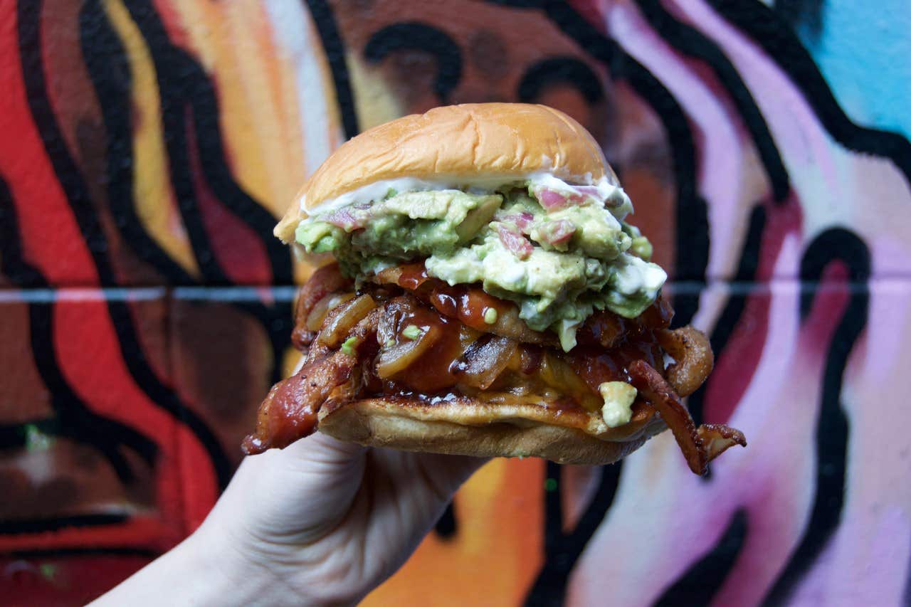 Hand holding a burger with bacon, guacamole, and sauce in front of a colorful graffiti background.