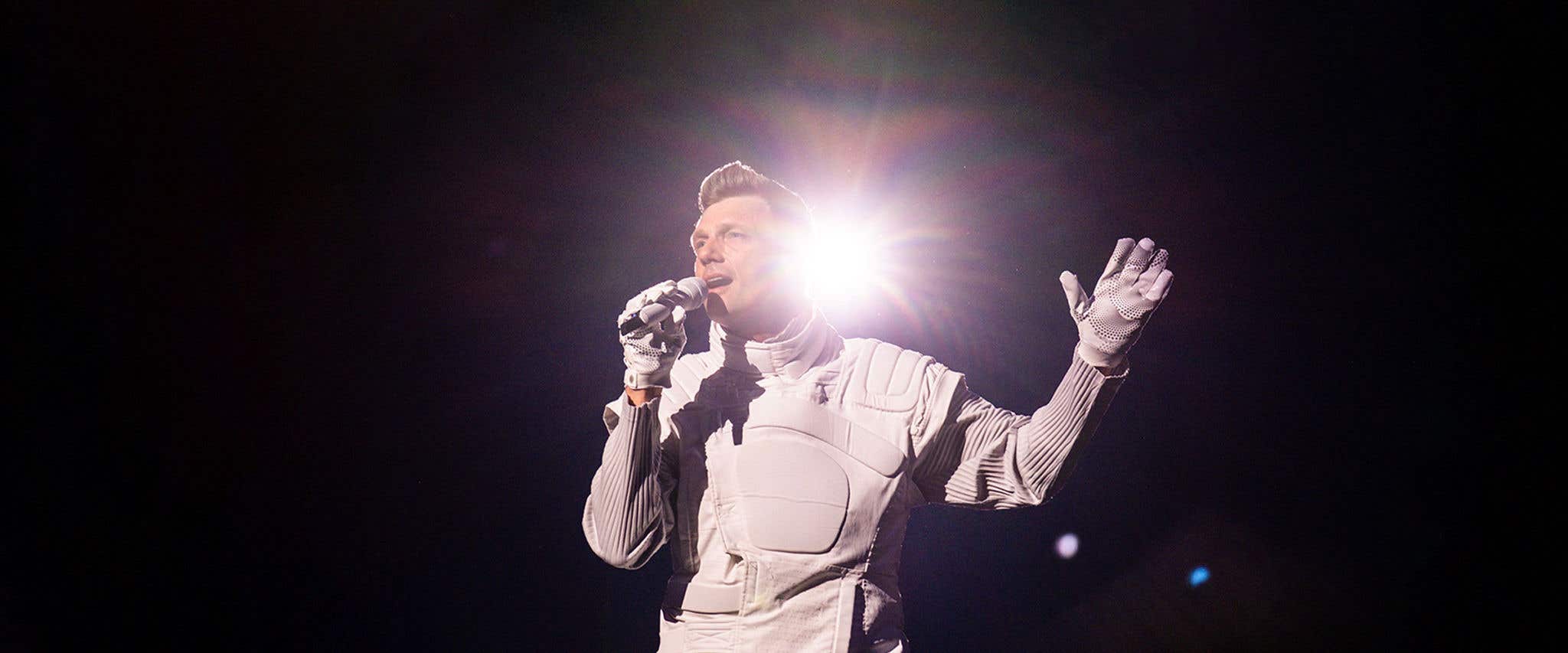 Nick Carter dressed in all white holding a microphone in front of a dark background with light shining from behind him.