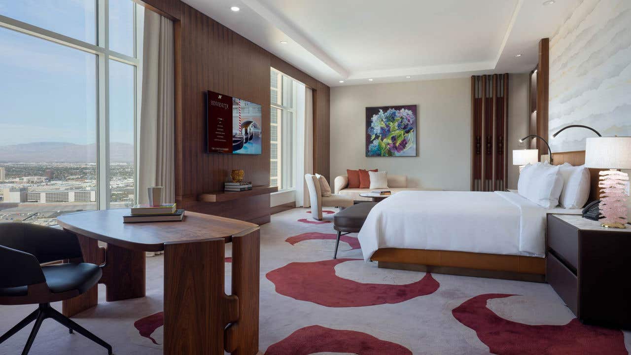 A large hotel bedroom with a king bed, desk, seating area, TV, and two large windows with a view of the Las Vegas valley.