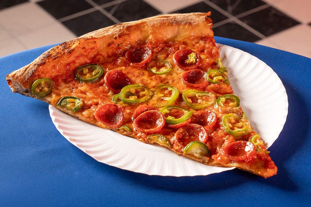 Large slice of pepperoni and jalapeno pizza sits on a white paper plate on a blue clothed table. 