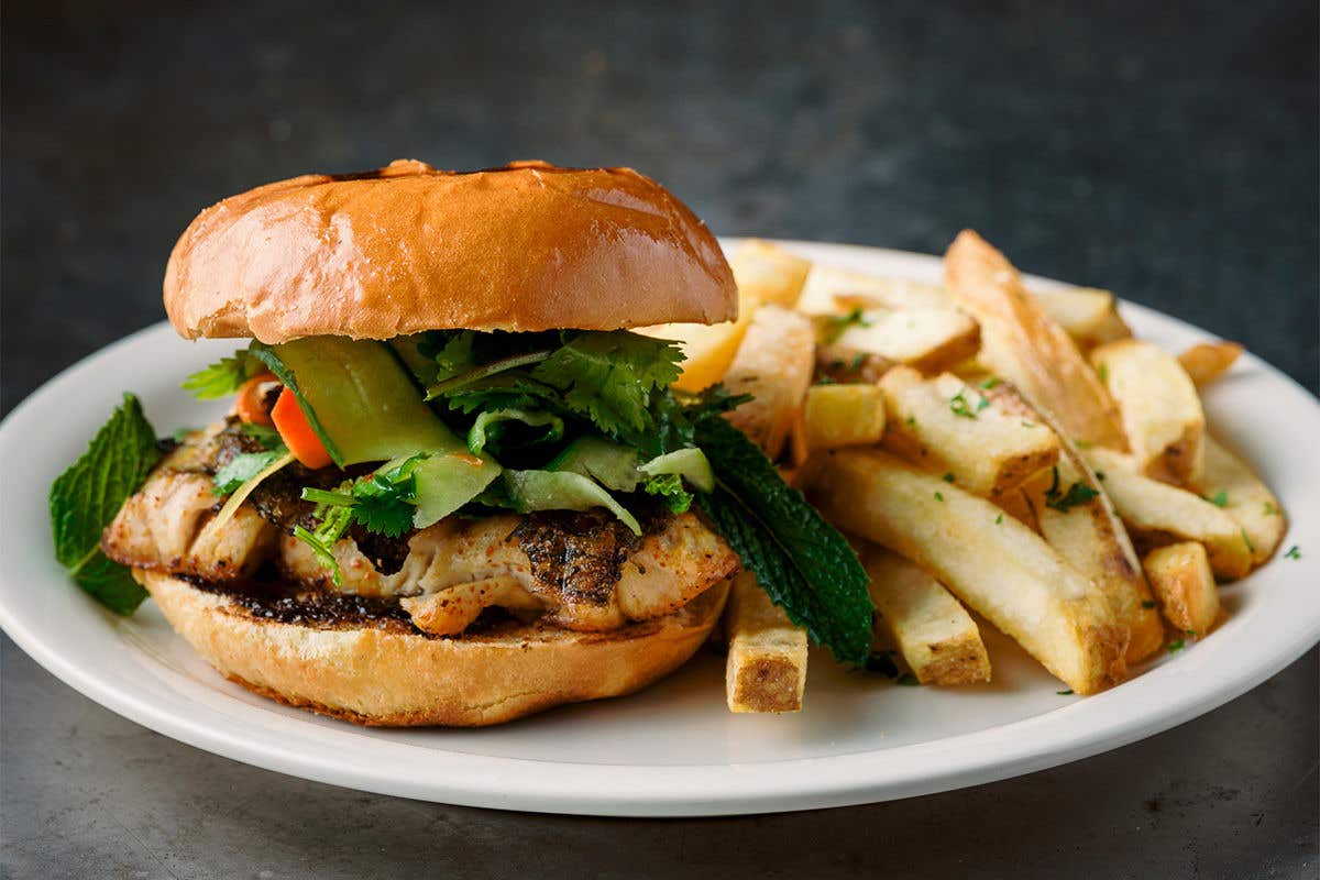 Blackened sea bass on a round bun topped with vegetables inside next to a pile of French fries. 