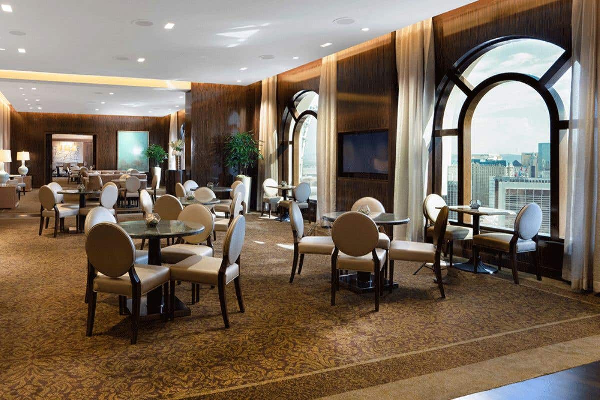 Spacious lounge with elegant decor, round tables, arched windows, a bar counter, and a person standing near the bar.