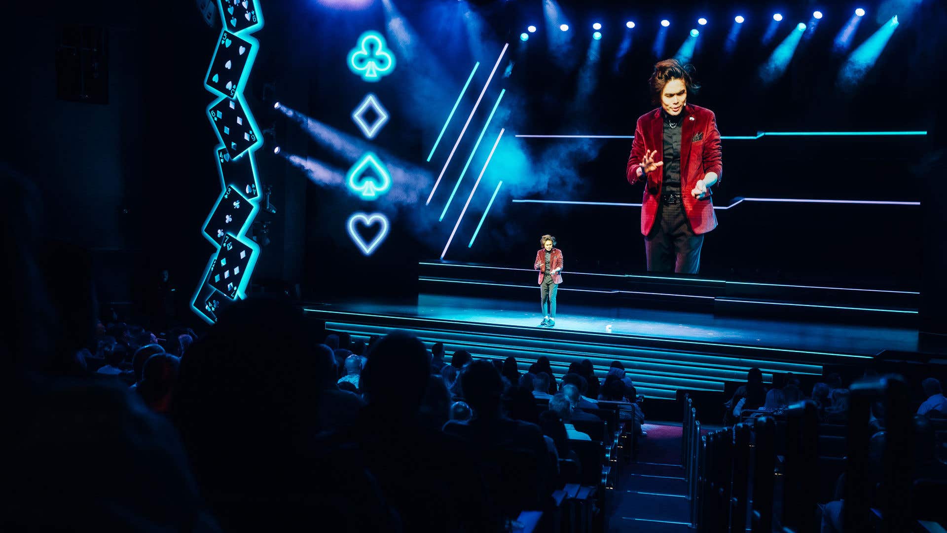 Shin Lim on stage wearing black shirt, pants, shoes, and a burgundy jacket with bigger projected image of the stage on the screen behind him. 