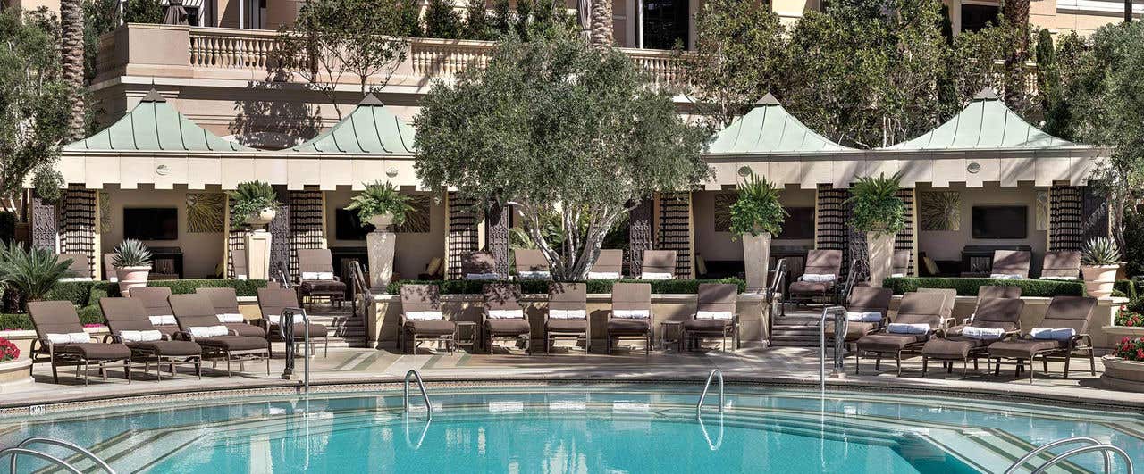 A pool with cabanas, lounge chairs and trees.