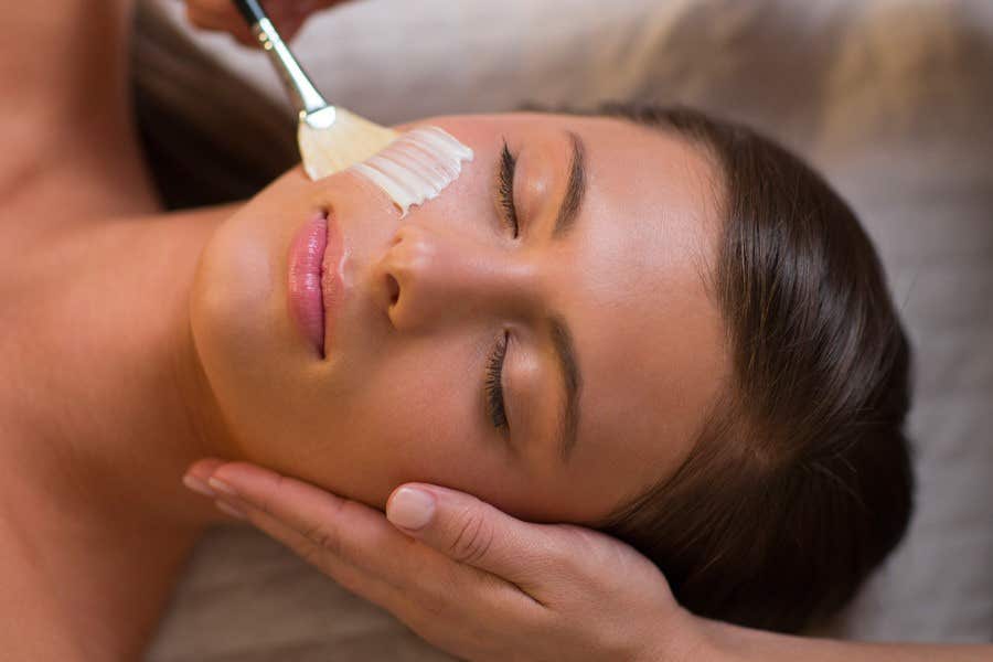 A person lying down with eyes closed while a facial mask is being applied with a brush.