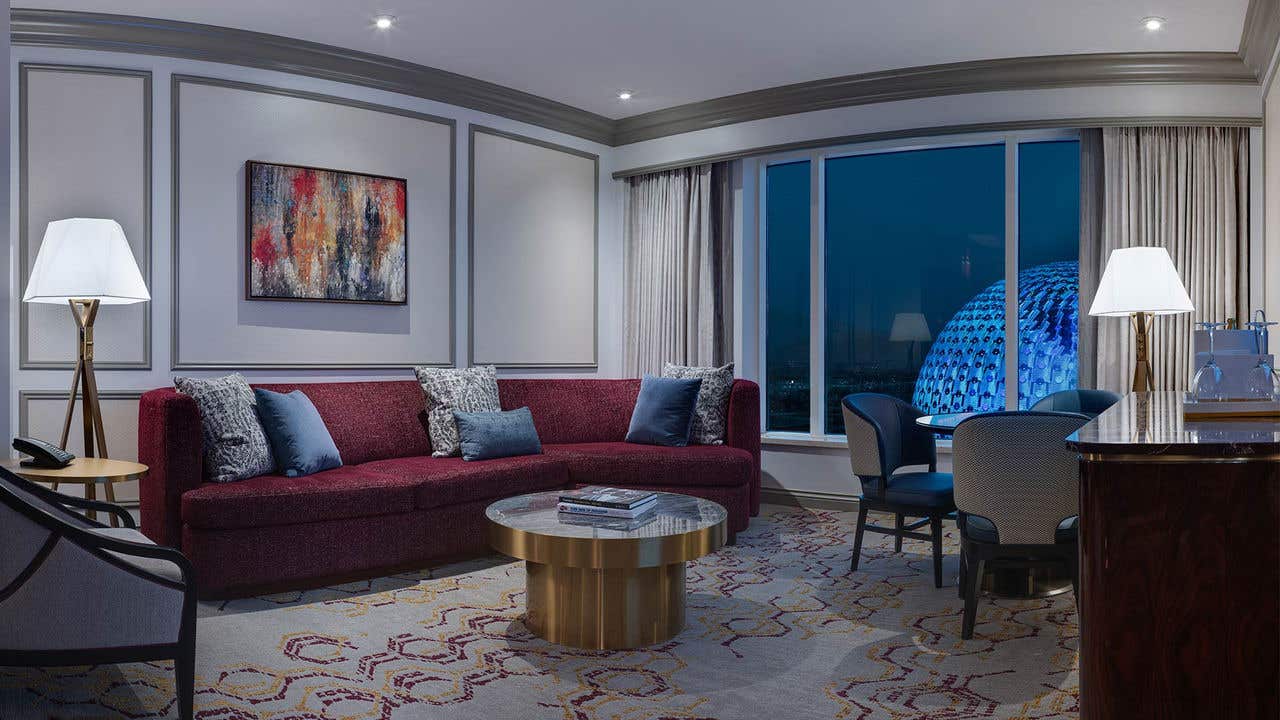 Living room of a hotel suite, at night. A red couch and gold table are visible and outside the window, Sphere glows. 