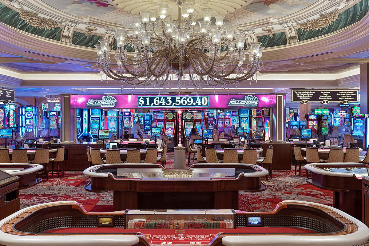 The Venetian Casino floor with some tables at the front, and some slots in the background, there is also an overhead chandelier above the tables
