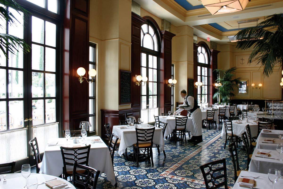 A waiter sets tables in an elegant, empty restaurant with large windows, white tablecloths, and patterned tile floor.