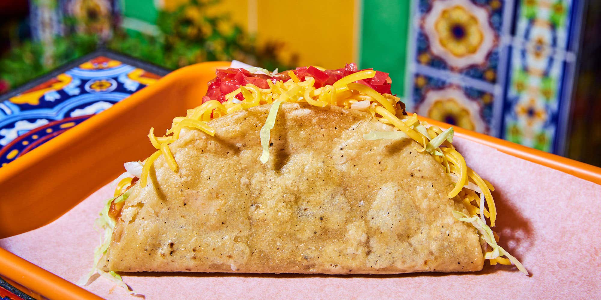 Large hard-shell taco with cheese and tomatoes on a orange tray in front of a colorful wall.
