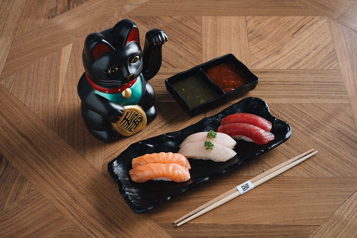 A black plate containing assorted types of sushi on it, alongside chopsticks, sauce trays, and a cat figurine, all on a brown wooden table