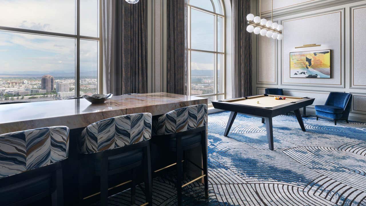 A hotel living room with a high-top table with six chairs, a pool table, two armchairs, and two tall arched windows with a view of the Las Vegas valley.