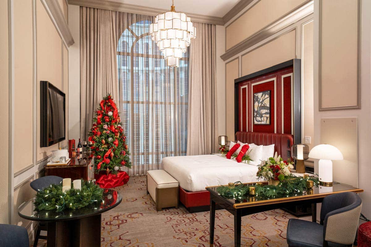 A hotel room with a king bed decorated for Christmas with a Christmas tree in the corner with red decorations, a garland on the desk next to the bed, a wreath with candles on the dining room table, candy and wine on the credenza, and two red stockings on the bed.
