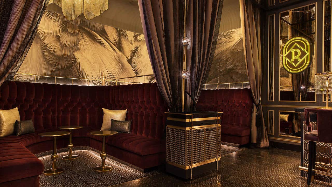 A gold toned bar with intricate floor, deep burgundy velvet booths and sparkling chandelier.