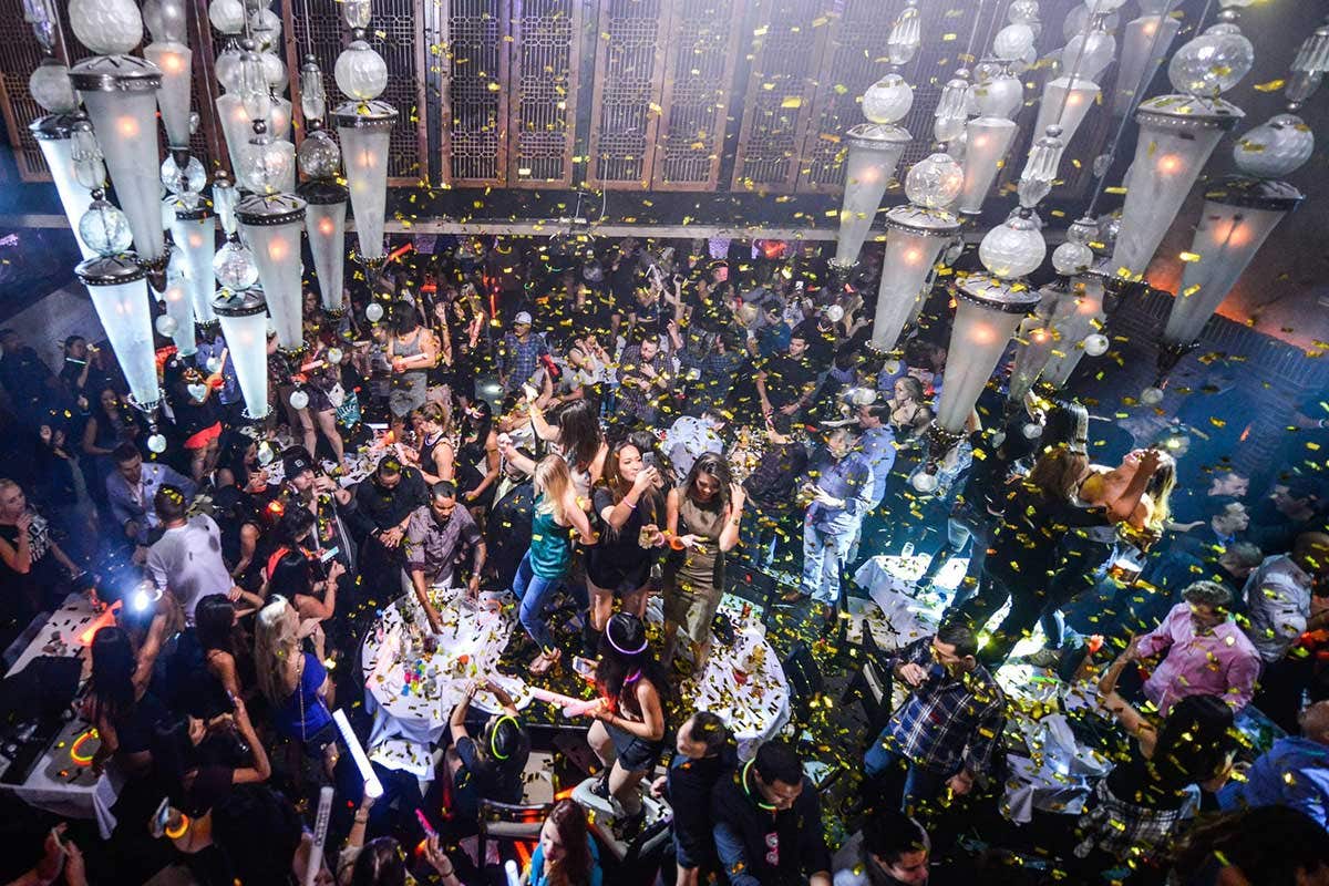 A crowded nightclub with people dancing, confetti falling, and vibrant lights hanging from the ceiling.