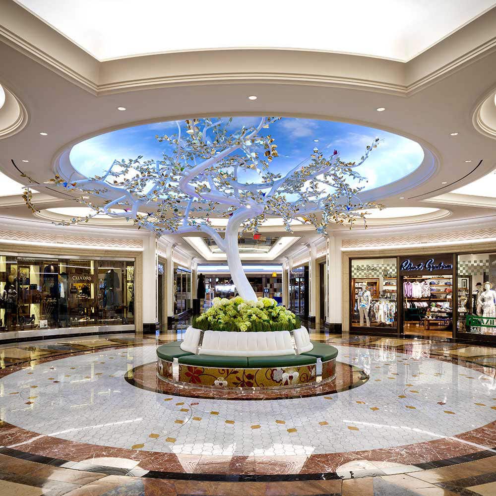 A luxurious indoor shopping area with a white tree sculpture and circular seating under a domed ceiling with a sky mural.