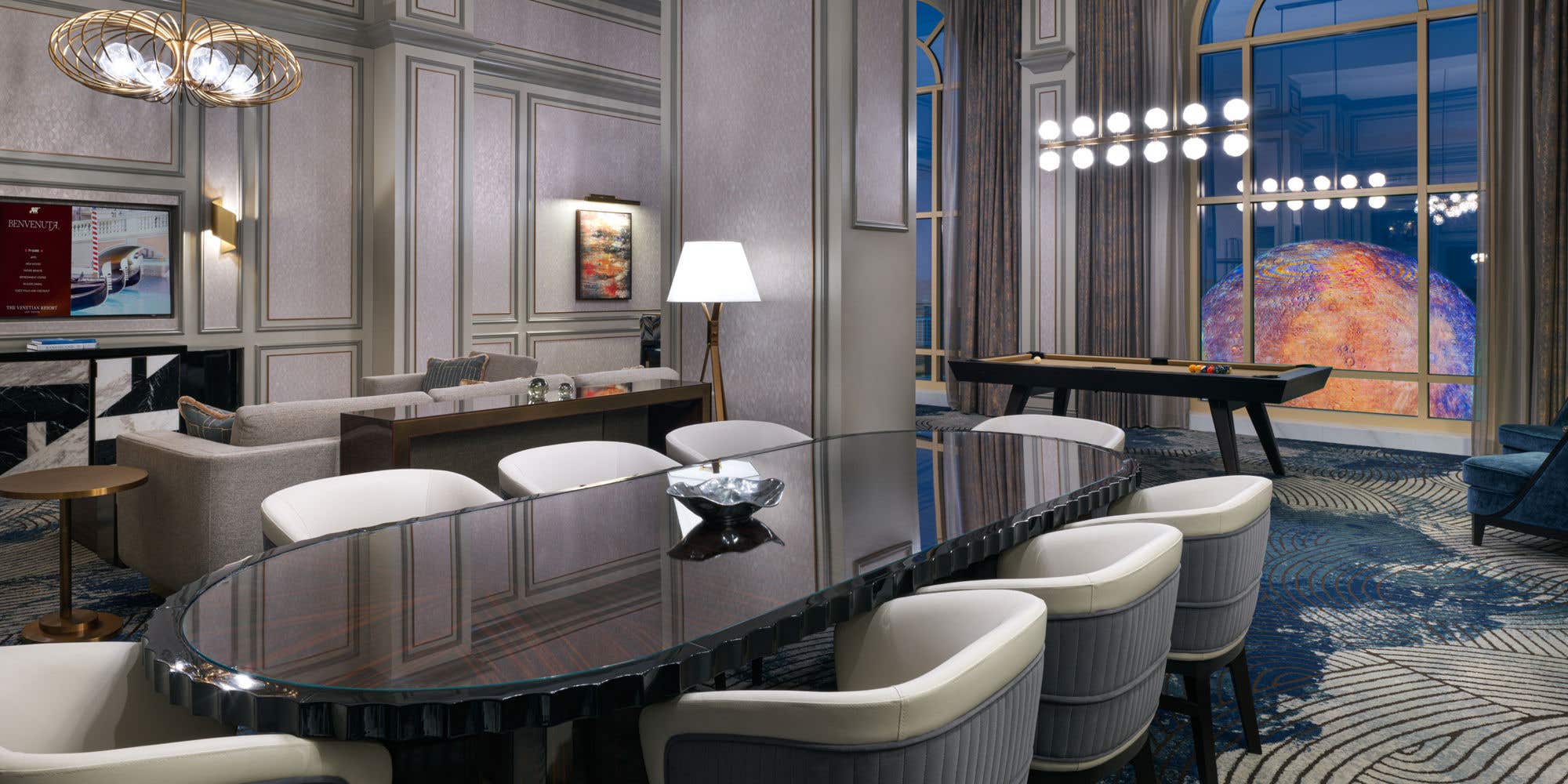 A modern luxury Corner Two Bedroom Suite living room featuring a long dining table, comfortable seating area, pool table, and large windows with nighttime views of the brightly lit Sphere.