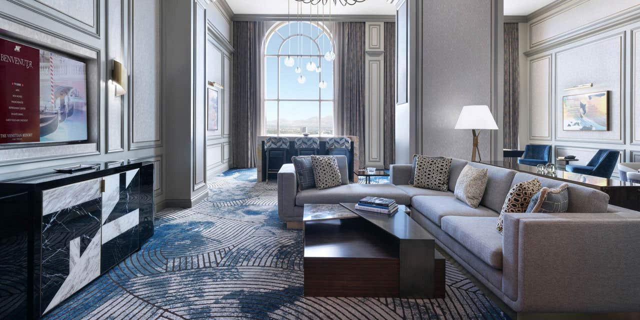 A hotel living room with a couch, coffee table, TV, a high-top table with bar chairs, chandeliers, armchairs, high ceilings, a tall arched window, and the edges of a pool table and dining table visible to the right.