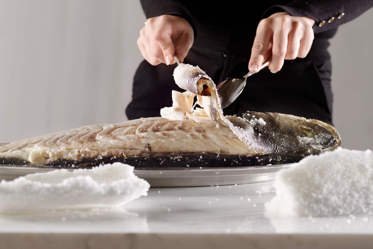 A person is cutting into a salt-baked fish on a silver tray with a fork and spoon.